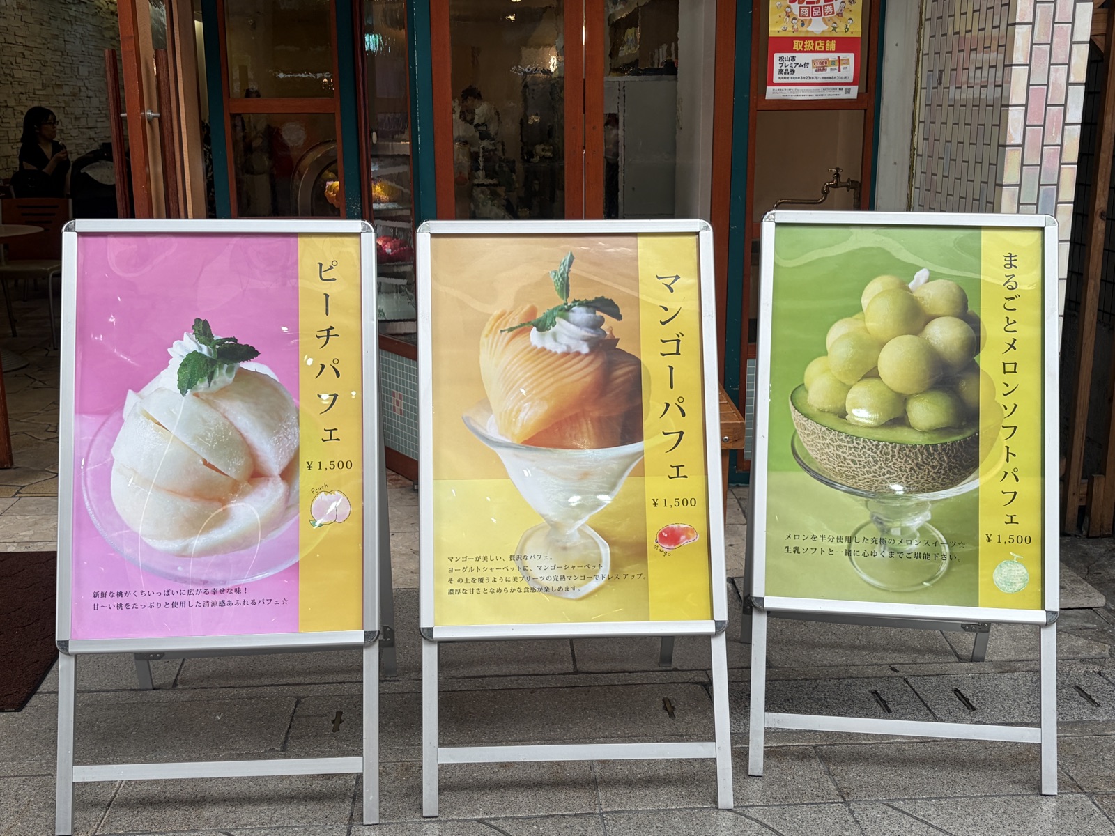 フルーツパーラーみしまの店頭に並ぶ、季節のパフェの看板。ピーチパフェ、マンゴーパフェ、まるごとメロンソフトパフェ