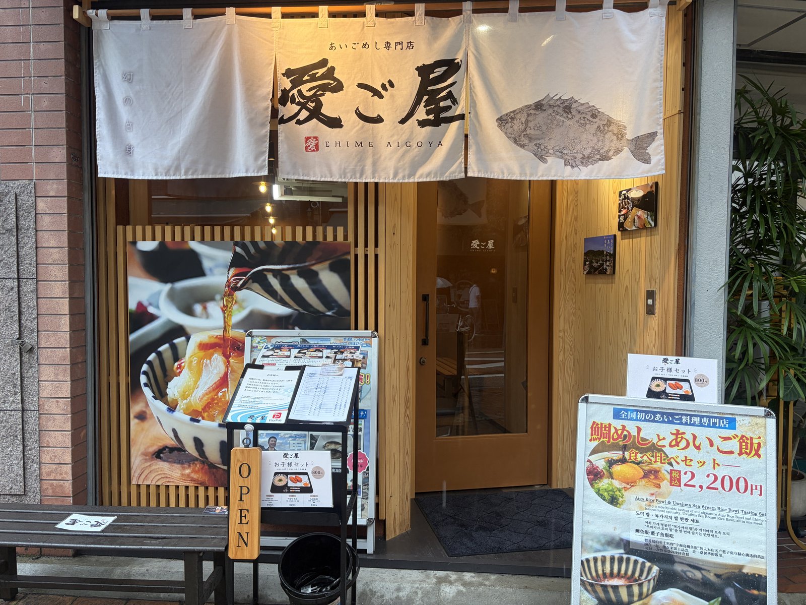 「愛ご屋」の文字と魚の絵が入った大きな暖簾。店先には「鯛めし・あいご飯 食べ比べ2,200円」の看板